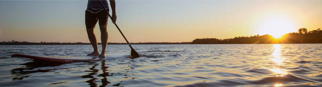 A man paddle boards across a calm lake as the sun over the trees in the distance.