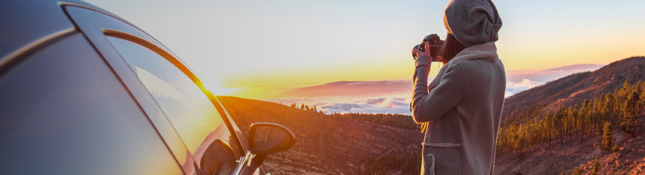 A woman standing next her car, looks out over the horizon and takes a picture of the sunset.