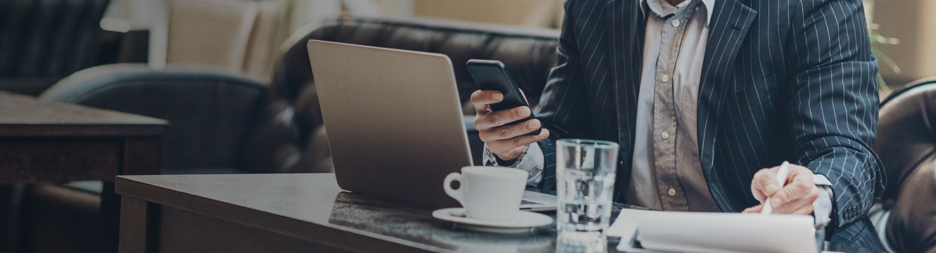 A business man sits at a table with his laptop and papers as he looks at his phone.